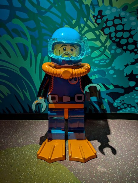 A LEGO diver figure wearing a blue helmet and orange fins against a tropical background at the SeaLife Centre in Malaysia.